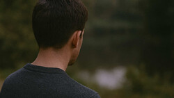 Man viewed from behind wearing a dark sweater, gazing toward blurred natural scenery.