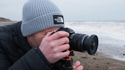 Photographer in winter beanie holding DSLR camera on beach with tripod and gimbal stabilizer.
