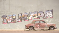Vintage red car parked in front of weathered wall mural spelling 'Seligman' with Route 66 imagery.