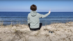 Person sitting on beach dunes facing ocean with one arm raised, wearing light hoodie and dark pants.