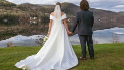 Bride and groom holding hands facing a lake with mountains and forested hills reflected in still water.