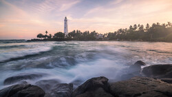 Lighthouse on a peninsula with palm trees, photographed from a rocky beach at golden hour with waves in the foreground.