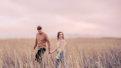 Two people walking through a dried grass field at dusk under a pink-toned sky.
