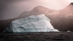 Massive tabular iceberg floating in dark water with snow-capped mountains and soft golden sunlight in the background.