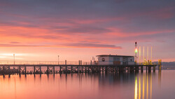 Wooden pier extending into still water at sunset with warm pink and orange sky reflecting on the surface.