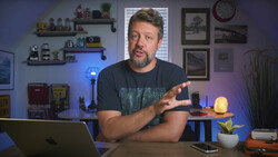 Man in dark t-shirt speaking to camera at desk with laptop and studio lighting setup visible.