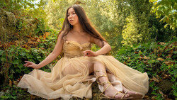 Woman in flowing beige tulle gown posed seated in wooded garden setting.