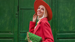 Woman in red coat and hat poses against a green door, holding a quilted green handbag.