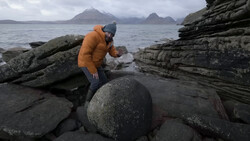 Person in orange jacket exploring rocky coastal landscape with layered basalt formations and mountains across the water.
