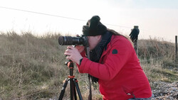 Photographer in red jacket looking through telephoto lens mounted on tripod in rural landscape.
