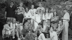 Black and white group portrait of approximately twenty people arranged in three rows outdoors among foliage.