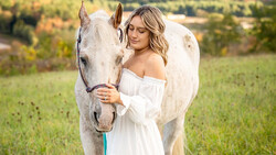Woman in off-shoulder white dress standing beside a gray horse in a pastoral field.