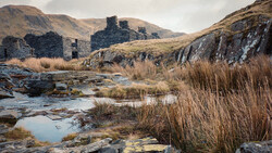 Ruined stone buildings in a moorland valley with a shallow stream and reeds in the foreground.