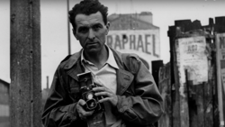 Man holding a vintage rangefinder camera in an urban setting with weathered buildings behind him.