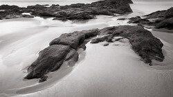 Rocky outcrops emerging from smooth sand on a misty beach with long-exposure water blur.