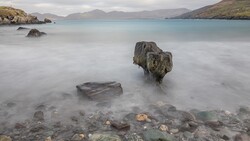 Moss-covered rock jutting from milky seawater with mountains across a bay under overcast sky.
