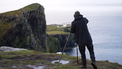 Hiker with trekking poles standing on mossy cliff edge overlooking ocean and coastal landscape.