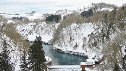 Snow-covered mountain landscape with a turquoise river flowing through a valley lined with evergreen and bare deciduous trees.