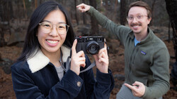 Woman holding a rangefinder camera while smiling at the camera, with a man gesturing enthusiastically in the background outdoors.