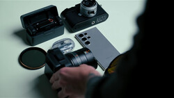Photographer's hands holding vintage film camera lens with smartphone, filters, and camera gear arranged on white surface.