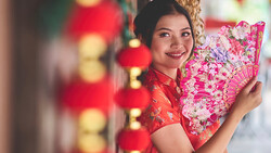 Woman in red traditional dress holding ornate pink fan amid festive lantern decorations.