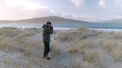 Photographer in dark jacket holding camera to face on sandy beach with dune grass and mountain across bay.
