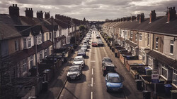 Overhead view of a residential street lined with parked cars and terraced houses under overcast sky.