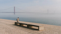 Solitary figure sitting on a bench facing a suspension bridge shrouded in fog.