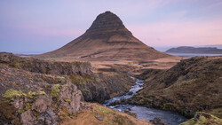 Kirkjufell mountain with snow-dusted slopes rising above a braided river valley at dusk.