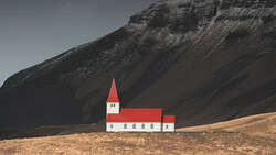 Red and white church with steeple standing alone on barren brown ground beneath a dark, textured mountain.