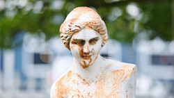 Weathered stone bust sculpture with patina and erosion marks, photographed outdoors against blurred background.