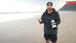 Photographer holding a telephoto lens on a misty beach while gesturing during discussion.