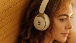 Woman wearing white over-ear headphones, photographed in profile against a warm wooden background.