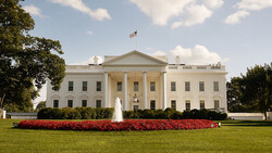 White neoclassical building with columned facade, manicured lawn, and red flowering plants in foreground.