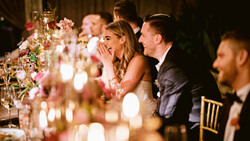 Bride and groom laughing together at an elegantly decorated wedding reception table with pink floral arrangements and candlelight.
