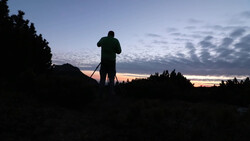 Silhouetted figure standing on elevated terrain with binoculars during twilight, surrounded by forested landscape.