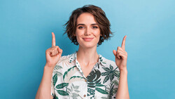 Woman with short dark hair wearing a tropical print shirt pointing upward with both index fingers against a blue background.