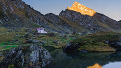 Alpine valley landscape with a pink building nestled between rocky peaks and a reflective pond in the foreground.