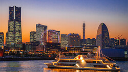 Illuminated ferry boat on water at dusk with city skyline and glowing buildings in background.
