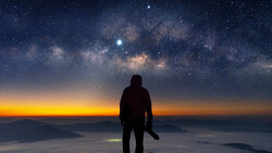 Silhouetted figure standing on a mountain peak facing the Milky Way galaxy at sunrise.
