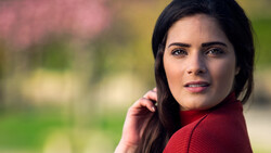 Woman with dark hair wearing a red sweater, looking at camera with soft expression against blurred background.