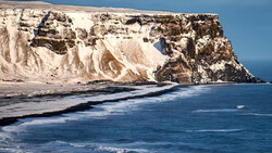 Dramatic white chalk cliffs with dark rock formations meet turquoise ocean waves under clear blue sky.