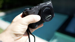 Hand holding a Canon compact camera with lens extended, displayed outdoors with blurred blue background.