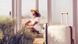 Woman in white dress and straw hat sitting by large windows with lavender plants and luggage beside her.