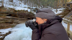 Photographer holding a camera to their face while standing in a snowy forest landscape.