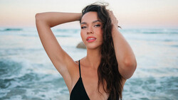 Woman in black swimwear posing at the ocean shore during golden hour sunset.