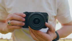 Photographer holding a compact mirrorless camera with a wide-angle lens, wearing a cream-colored shirt and gold ring.