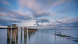 Weathered wooden pilings extend into calm water at dusk under a dramatic cloudy sky.