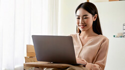 Woman in beige cardigan smiling while working on laptop at desk with cardboard boxes.