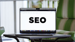 Laptop displaying the word SEO on its screen, positioned on a desk with green striped upholstered furniture visible in the background.